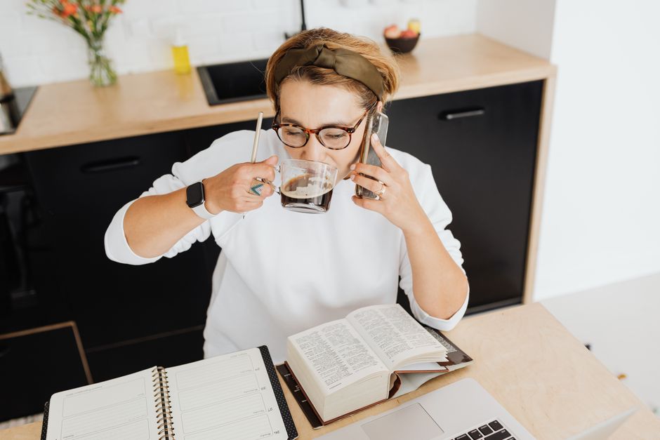 Adult woman drinking coffee, using smartphone, and studying at a kitchen table.
